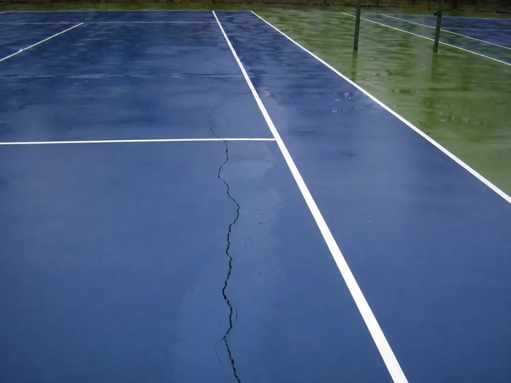 Cracked tennis court surface with visible line damage and water on outdoor court